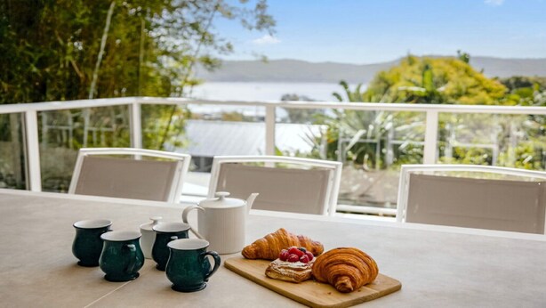Una mesa con un croissant y bayas, una tetera y cuatro tazas verdes en una terraza con vistas al agua y follaje.