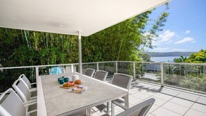 A modern outdoor dining area with a table set for two, surrounded by chairs, overlooking a lush garden and a scenic view of the sea. - Heated Plunge Pool with Bay Views! (WAGSTAFFE)