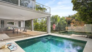 Una casa moderna con una gran piscina, una terraza de madera y una barandilla de cristal con vistas a un jardín.