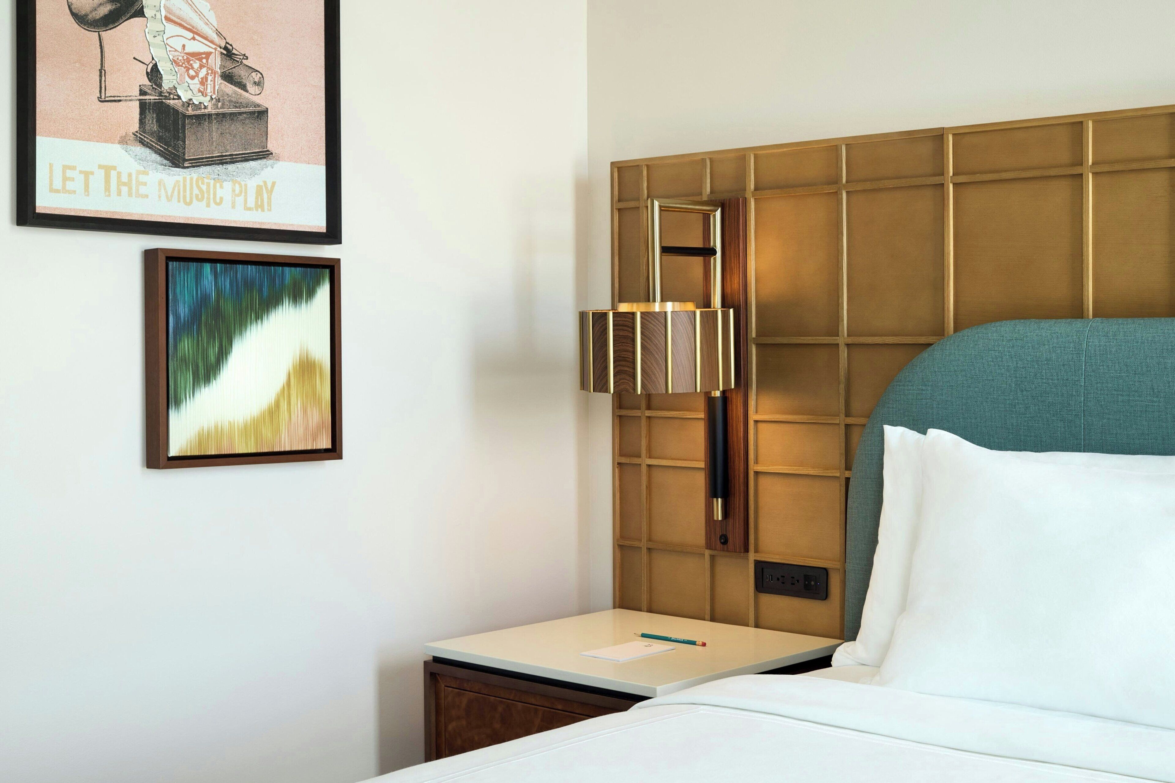 A hotel room with a bed, a nightstand, a wall-mounted lamp, and two framed pictures.