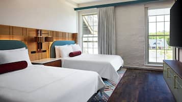 Habitación de hotel con dos camas, un ventanal grande con cortinas, un cabecero de madera y un televisor a la derecha.