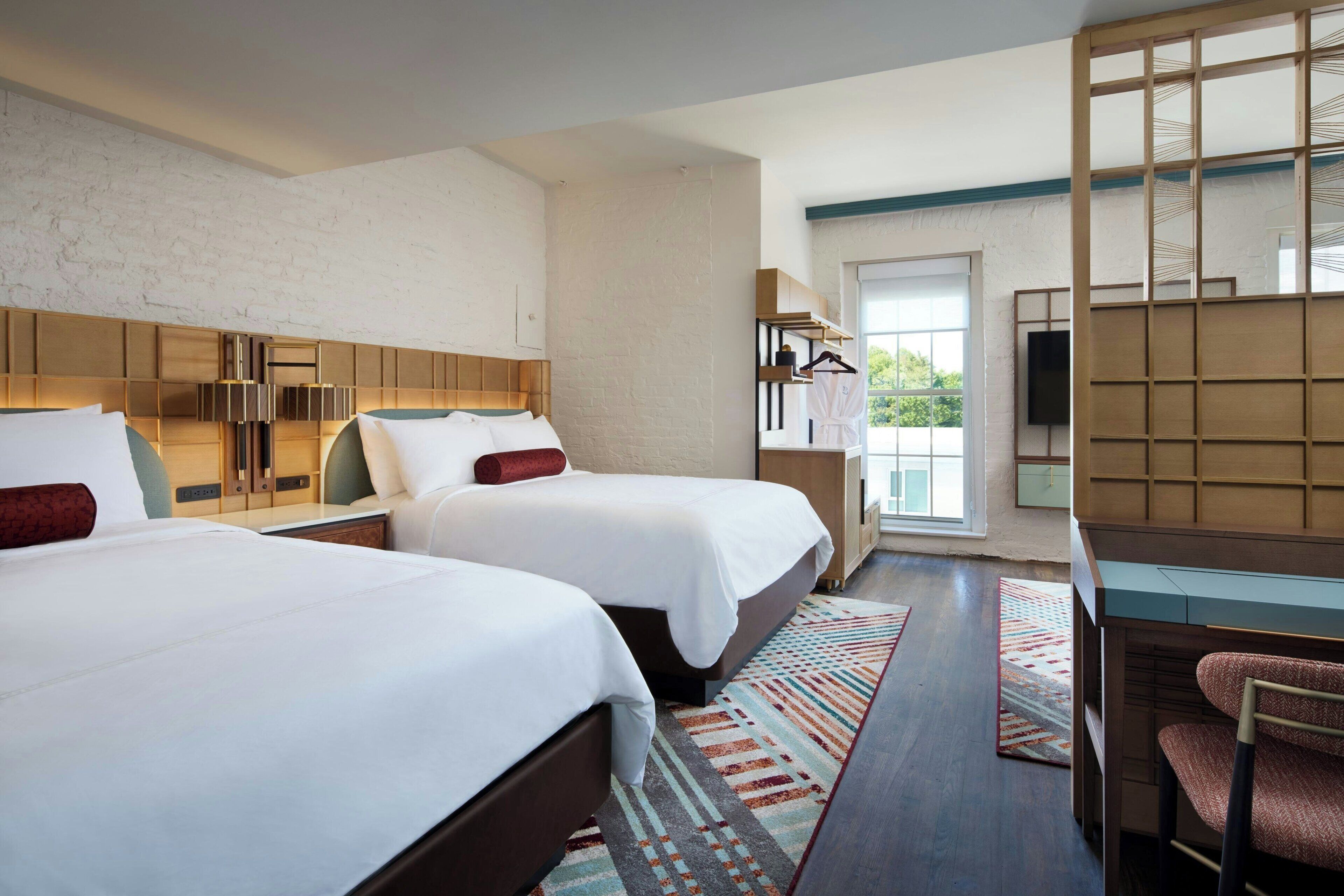 A hotel room with two beds, a desk, a chair, a TV, and a view of the outdoors.