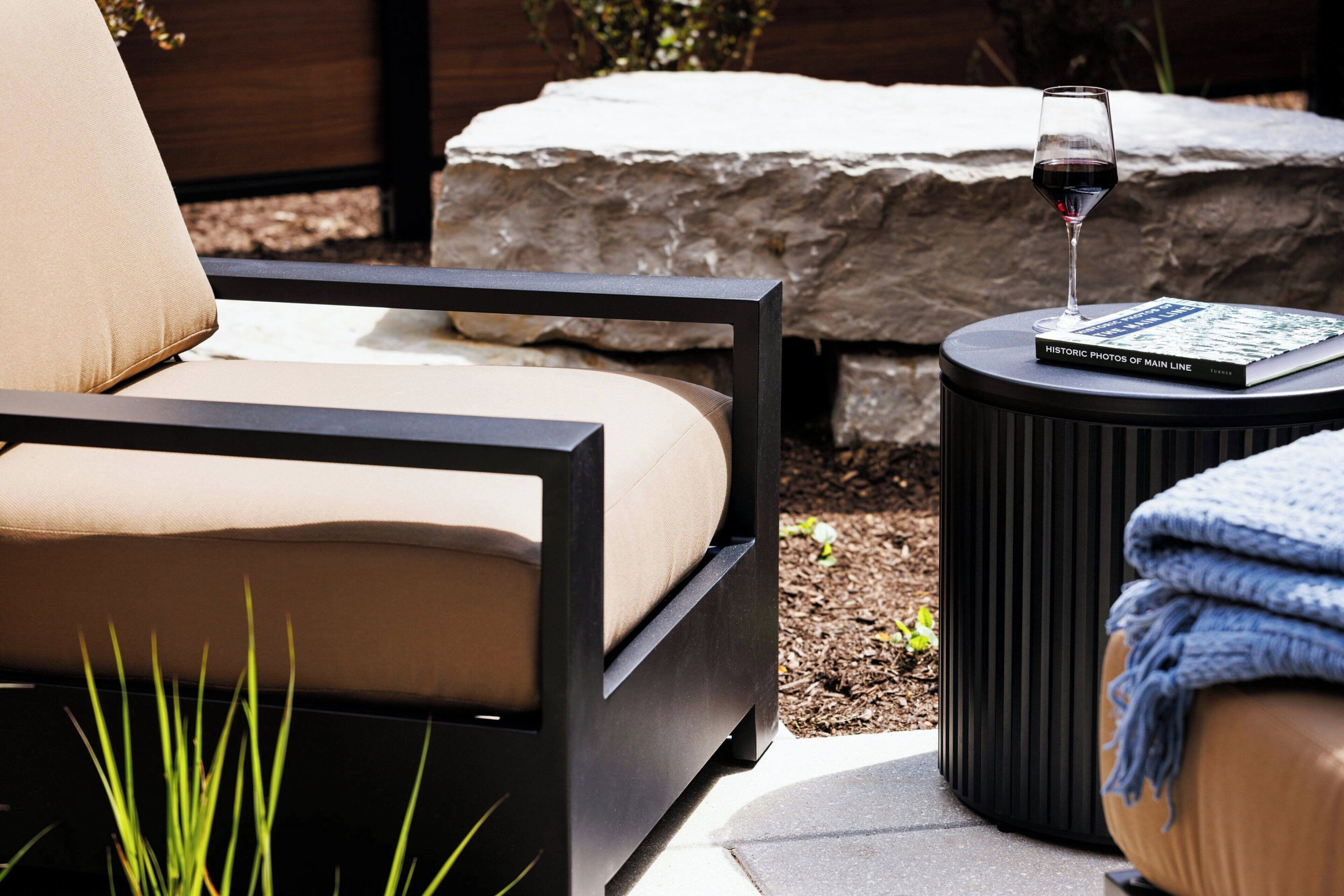 A modern outdoor seating area with a cushioned chair, a small round table with a book and a glass of red wine, and a stone backdrop.