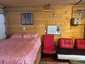 Una habitación de cabaña de madera con cama, una silla roja, un sofá blanco y negro, un equipo de aire acondicionado y una pared con un letrero que dice 'Old Guys Rules'.