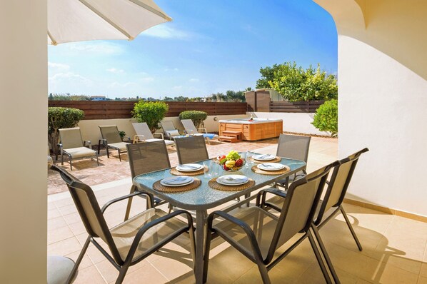 An outdoor patio with a table set for four, surrounded by chairs, and a hot tub in the background.