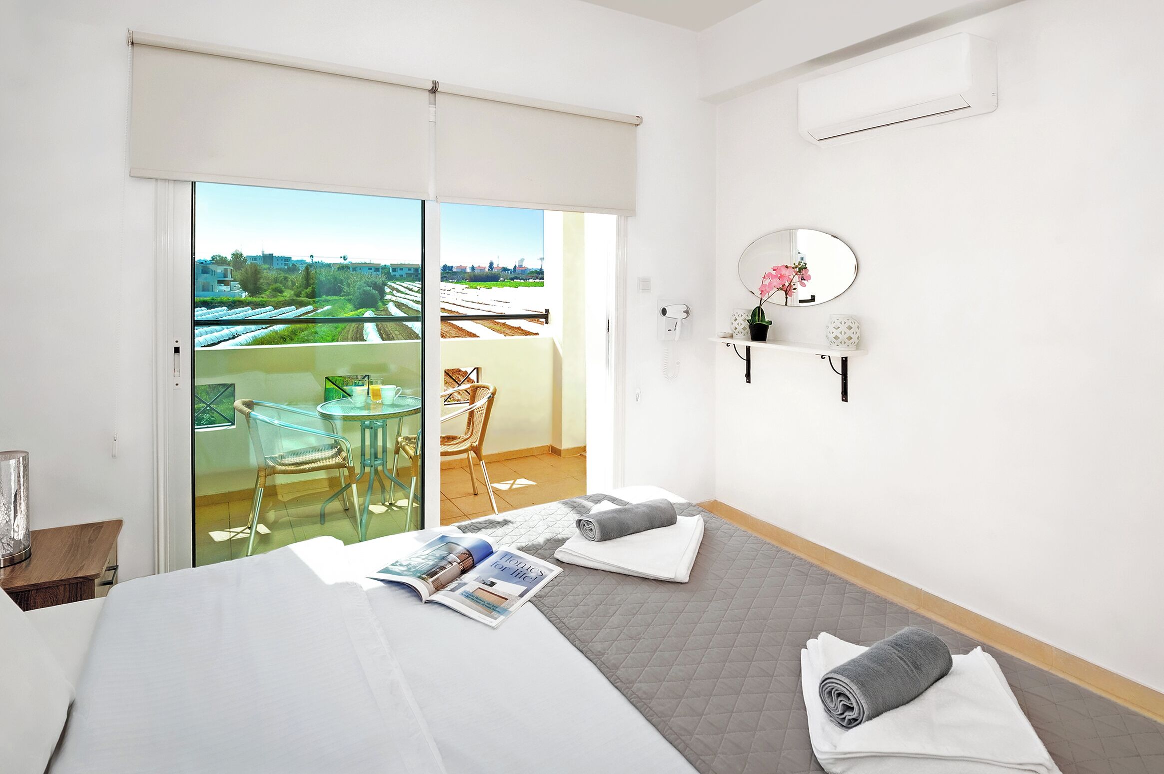 A hotel room with a bed, a magazine, a towel, a small table with a chair, a balcony with a glass door, and a view of greenery.