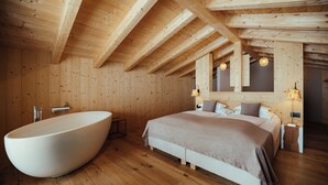 A wooden interior with a large bathtub, a bed with a beige bedspread, and a wooden ceiling with exposed beams. - Villa Flora Mountain Lodges (San Cassiano)