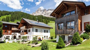 A mountain lodge with wooden architecture, a patio with lounge chairs, and a view of the mountains. - Villa Flora Mountain Lodges (San Cassiano)