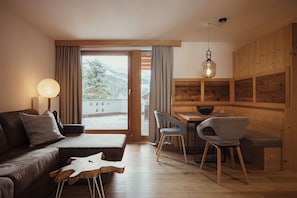 A modern living room with a sofa, chairs, a wooden dining table, and a view of the outdoors. - Villa Flora Mountain Lodges (San Cassiano)