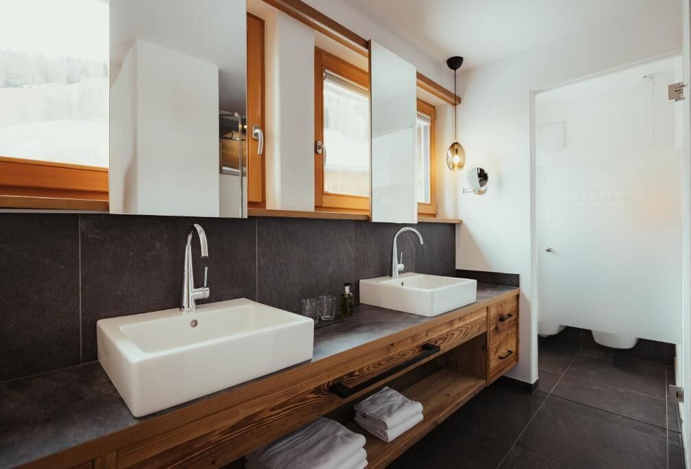 Ein modernes Badezimmer mit zwei Waschbecken, einem Holz-Waschtisch, dunklen Fliesen und einem großen Spiegel.