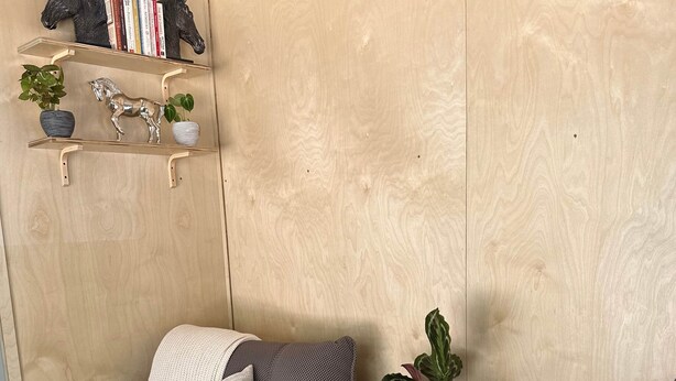 Un rincón acogedor con un sofá gris de estampado, una mesita redonda con un jarrón azul, y una pared de madera con estantes que exhiben libros y objetos decorativos.