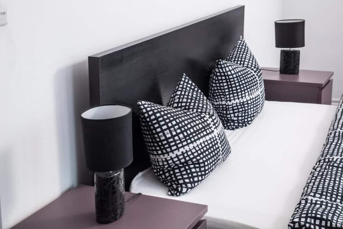 Uma cama com almofadas estampadas em preto e branco, uma luminária preta sobre uma mesinha de cabeceira e outra luminária preta em um criado-mudo.