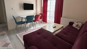 Un salon comprenant un grand canapé, une table à manger avec des chaises, un téléviseur à écran plat fixé au mur et une fenêtre ornée de rideaux rouges.