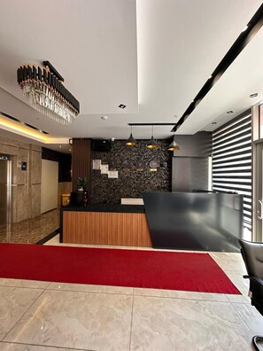 A modern reception area with a marble floor, a red carpet, a wooden reception desk, a black wall with clocks, and a hanging light fixture. - ada mevan otel (Van)