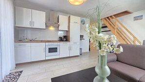 Una cocina moderna con armarios blancos, horno empotrado y cafetera. Se ve una escalera con barandillas de madera que sube a un nivel superior. En la zona de estar, hay un jarrón con flores blancas sobre una mesa.