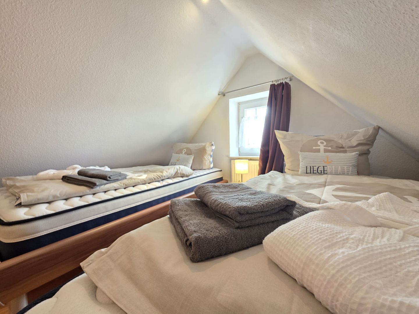 A cozy, well-lit attic bedroom with a bunk bed and a single bed, featuring a window with curtains, a lamp, and a visible bedspread.