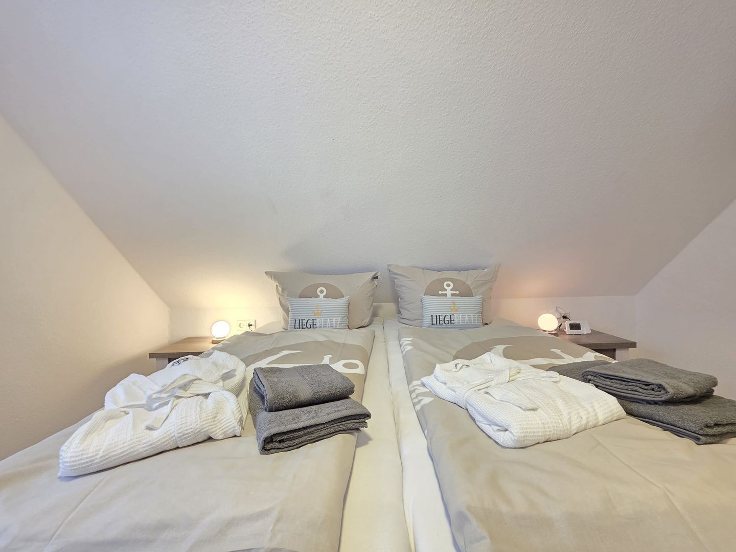 A twin bedroom with two beds, each with a pillow featuring a nautical anchor symbol, a bedside table with a lamp, and folded towels on the beds.