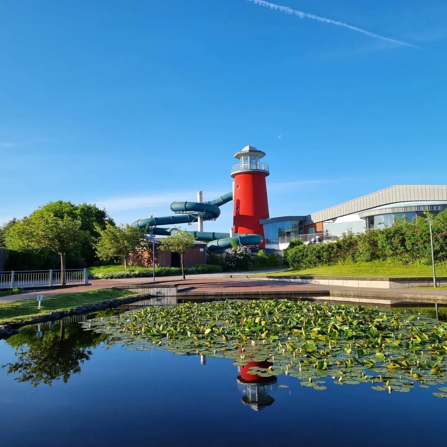 Un faro rojo con un elemento acuático en primer plano, rodeado de vegetación y un edificio moderno.