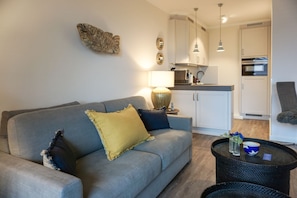 Ein Wohnzimmer mit grauem Sofa, einem gelben Kissen und einem schwarzen Couchtisch mit einer Vase und einer blauen Tasse. Im Hintergrund ist eine Küche mit weißen Schränken und einer Pendelleuchte erkennbar.