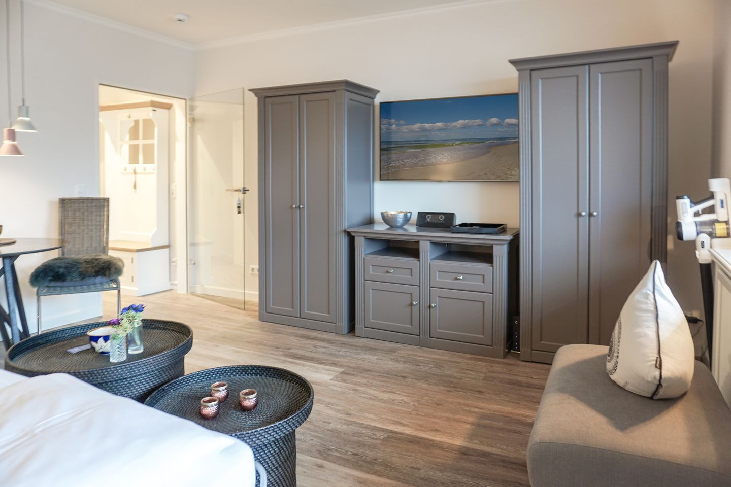 Hotelkamer met een bed, een zithoek met een stoel en een tafeltje, een grote grijze garderobe, een flatscreen-tv en een schilderij van een strand.