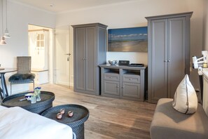 Ein Hotelzimmer mit Bett, einer Sitzecke mit Sessel und kleinem Tisch, einem großen grauen Kleiderschrank, einem Flachbildfernseher und einem Bild einer Strandansicht an der Wand.