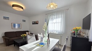 Ein modernes Wohnzimmer mit einem weißen Sofa, einem Esstisch mit Stühlen, einem Fernseher auf einer Kommode und einem Fenster mit durchsichtigen Vorhängen.