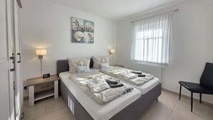 Ein Doppelbett mit passenden Kissen, ein Nachttisch, eine Lampe, ein Fenster mit Vorhängen und ein Sessel.