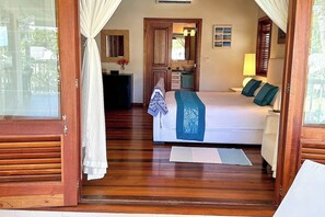A bedroom with a bed, a bedside table, a mirror, a window with curtains, and a wooden floor.