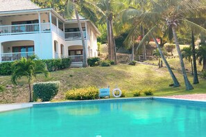 Un edificio de dos plantas con piscina, rodeado de palmeras y un espacio cubierto de césped.