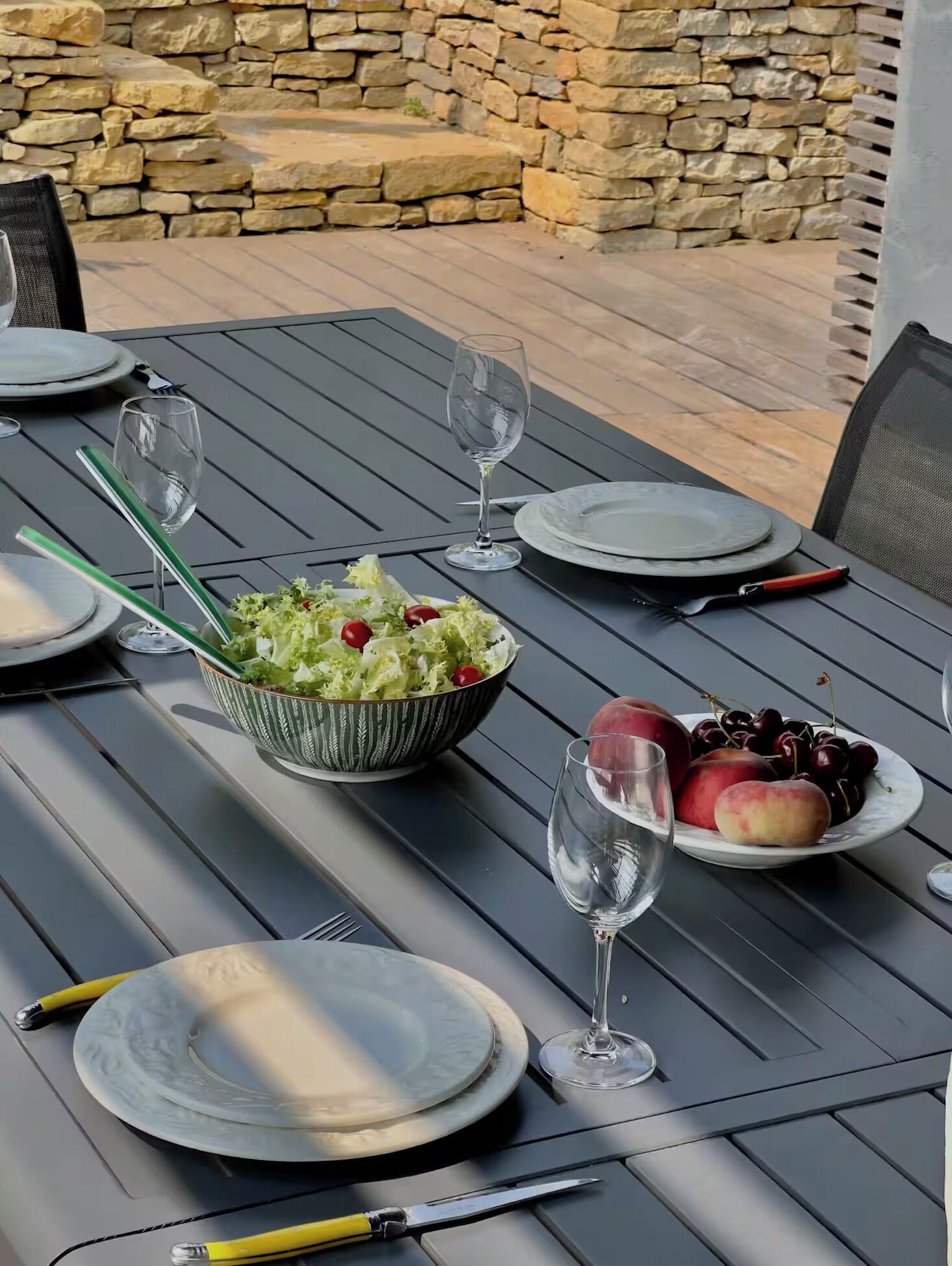 Un espacio de comedor al aire libre con un muro de piedra de fondo, con una mesa dispuesta con platos, vasos, cubiertos y un bol de ensalada y fruta.