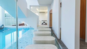 A modern indoor swimming pool area with a clear glass wall, a pool with a blue tint, and a staircase leading to a higher level. - The Pool Space — ICONIC Pool Villa (Pattaya)