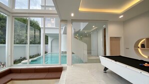 A modern interior with a pool, a billiards table, and a staircase leading to an upper level. - The Pool Space — ICONIC Pool Villa (Pattaya)