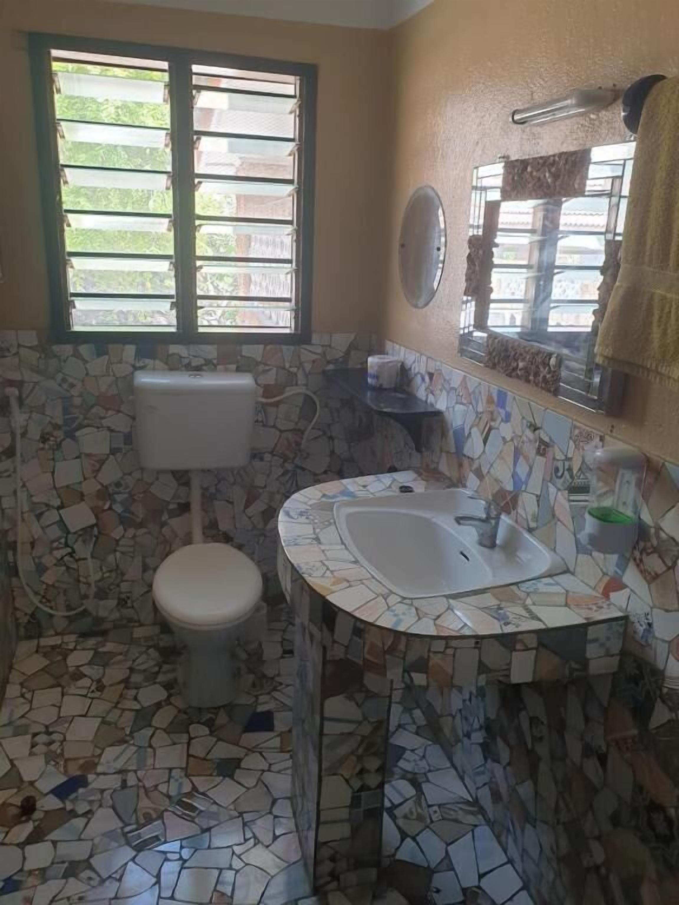 Une salle de bain avec un sol en mosaïque, des toilettes, un lavabo et une fenêtre avec des stores.