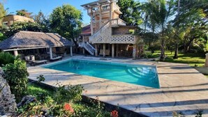 Una piscina con solárium de piedra, una estructura cubierta con techo de paja y un edificio de varios pisos con balcón.