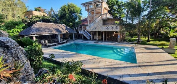 Una piscina con solárium de piedra, una estructura cubierta con techo de paja y un edificio de varios pisos con balcón.