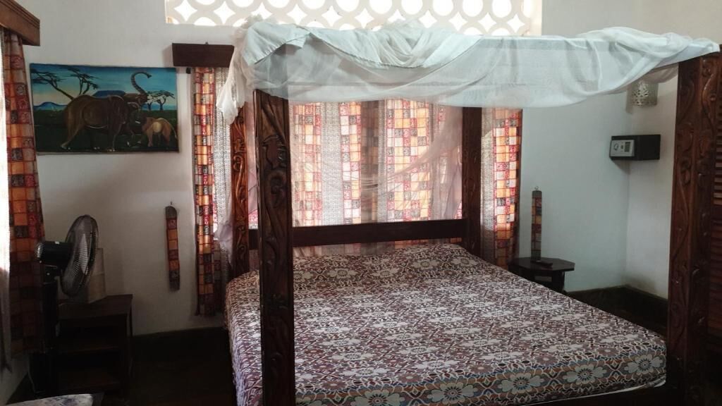 A room with a bed, curtains, a fan, a painting, and a window with a patterned view.