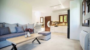 Een moderne woonkamer met een bank, salontafel en poef. Achtergrond toont een keuken met witte kasten en een raam.