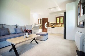 Un salon moderne avec un canapé, une table basse et un pouf. On aperçoit une cuisine aux meubles blancs et une fenêtre à l’arrière-plan.