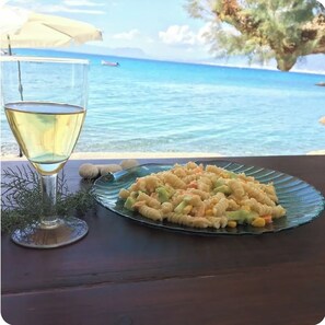 A glass of white wine and a plate of pasta with vegetables on a table by the sea. - Safe & Comfortable 1BR Seaside Apartment with A/C (KALAMATA)