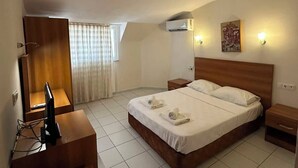 A hotel room with a bed, bedside tables, a desk with a computer, an air conditioning unit, and a window with curtains.