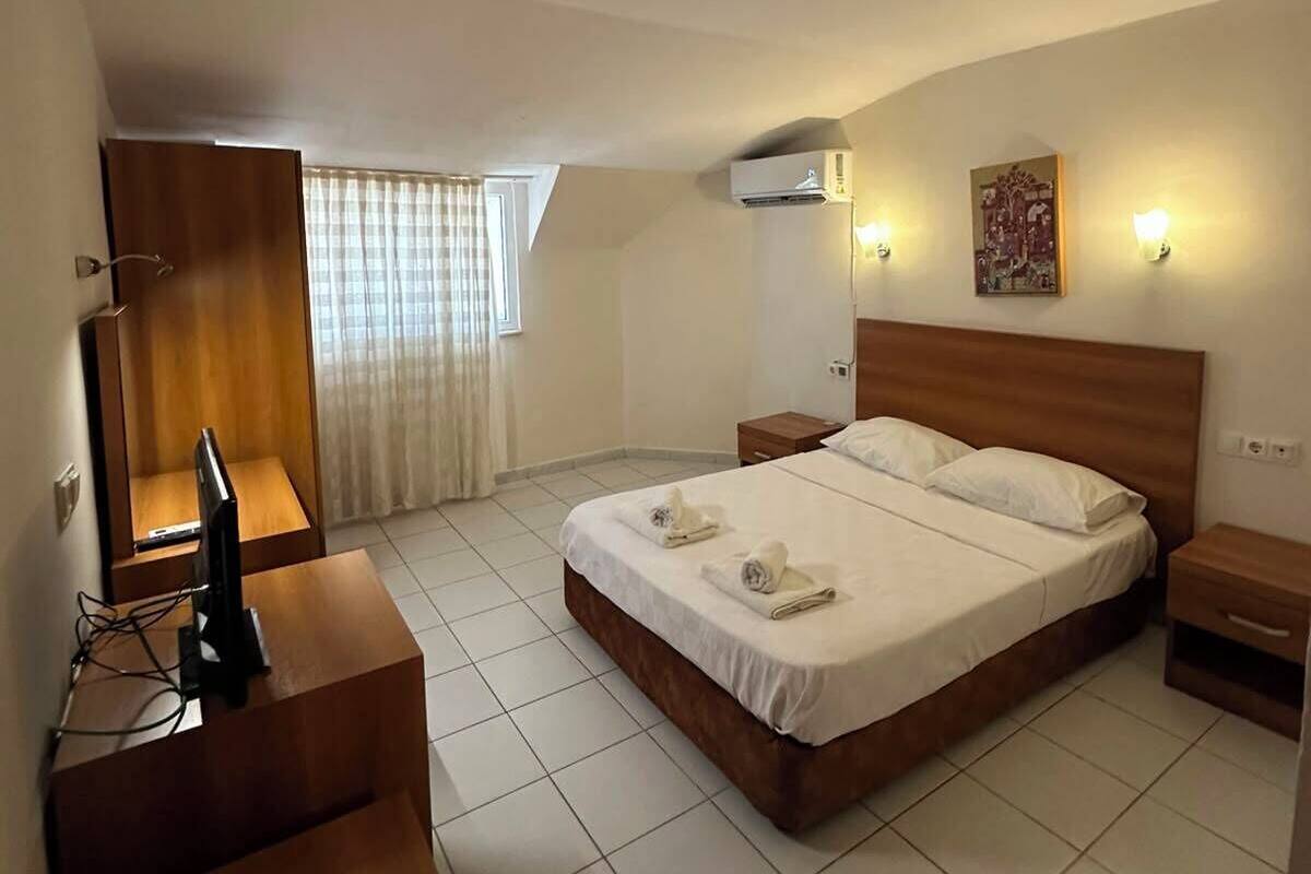 A hotel room with a bed, bedside tables, a desk with a computer, an air conditioning unit, and a window with curtains.