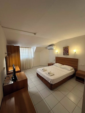 Une chambre d’hôtel comprenant un lit, des tables de chevet, un bureau avec un ordinateur, un appareil de climatisation et une fenêtre avec des rideaux.