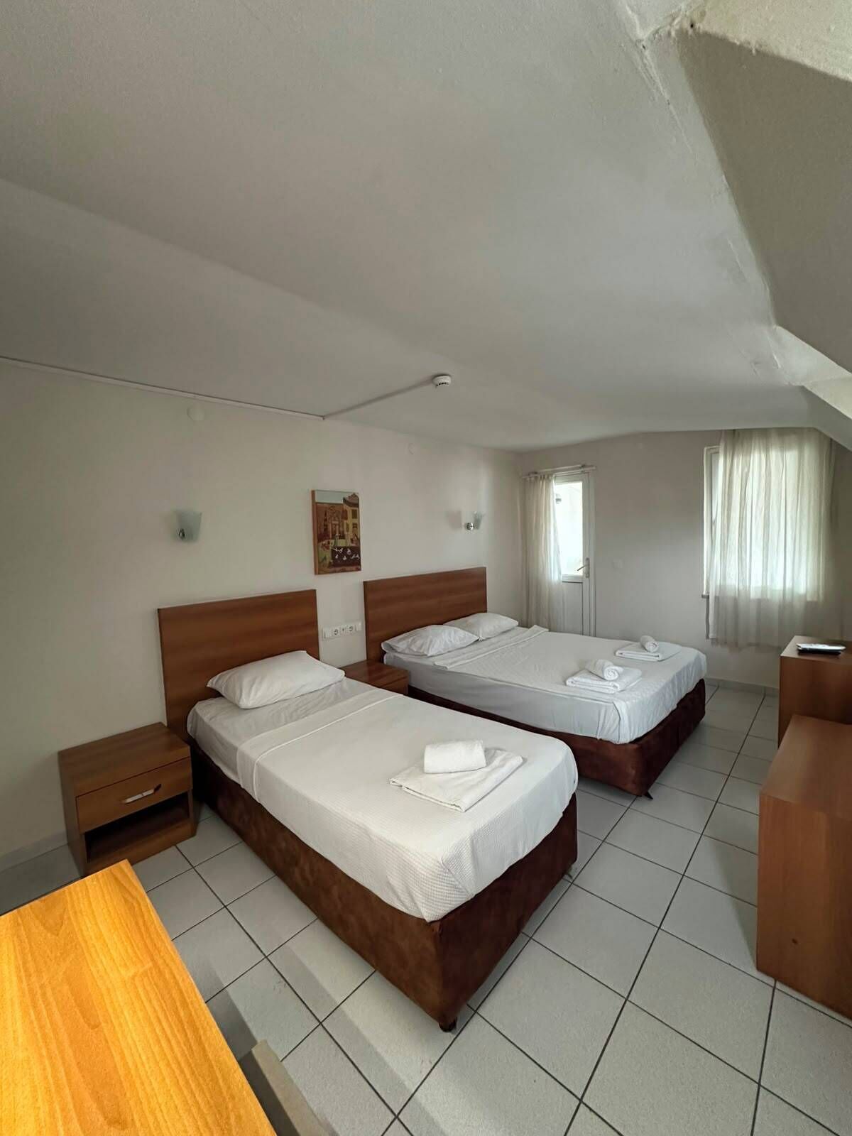 A hotel room with two beds, a wooden desk, a small bedside table, a wall-mounted light, and a window with curtains.