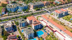An aerial view of a coastal resort area with multiple buildings, including a hotel with a swimming pool and a beach in the background.