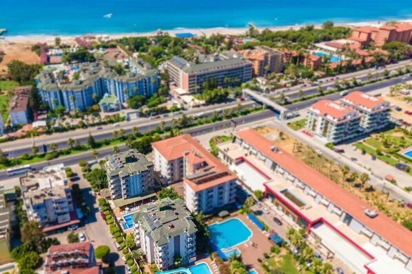 Vue aérienne d’une zone de villégiature côtière comprenant plusieurs bâtiments, dont un hôtel avec une piscine, et une plage en arrière-plan.