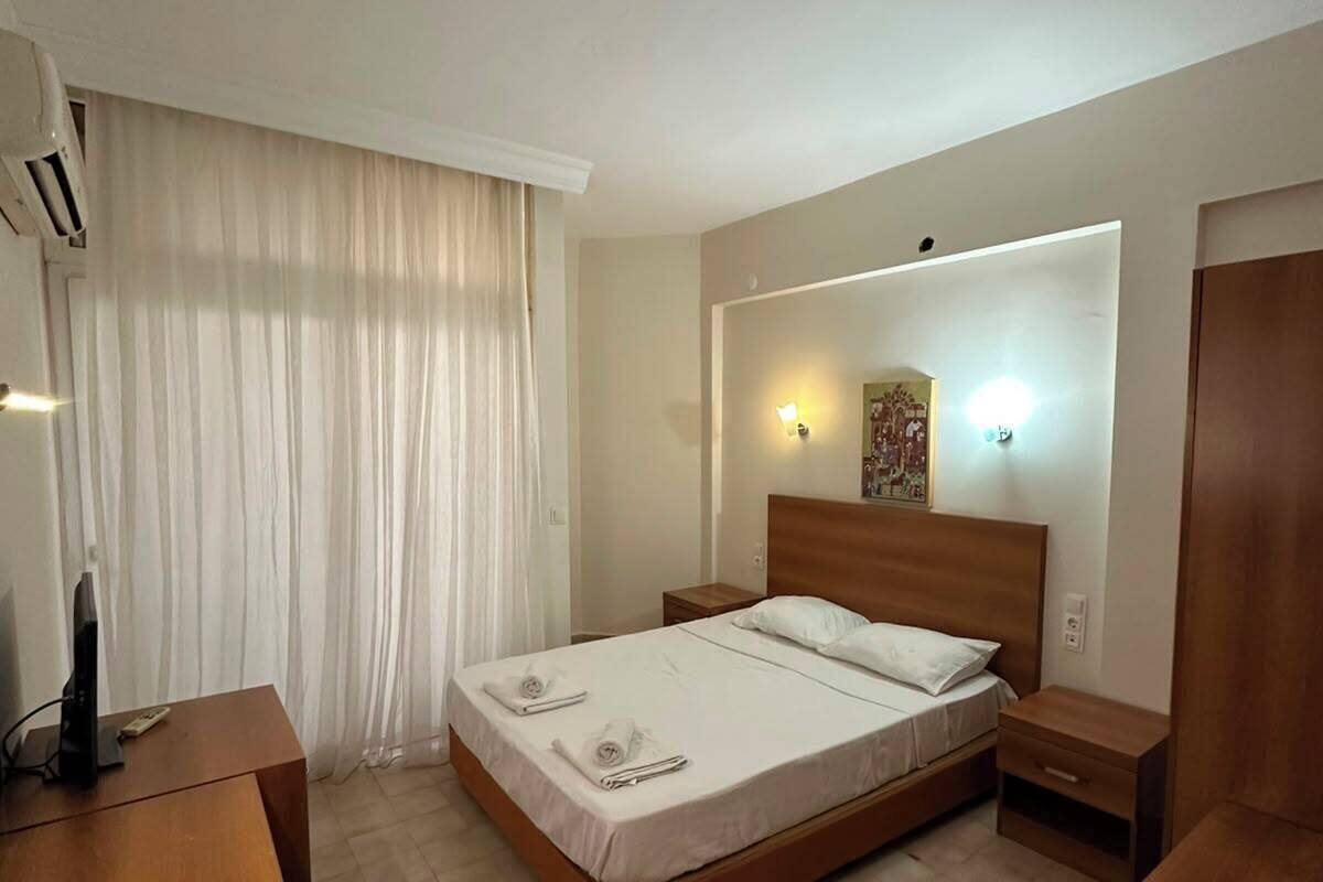 A hotel room with a bed, bedside tables, a desk, an air conditioning unit, and a window with curtains.