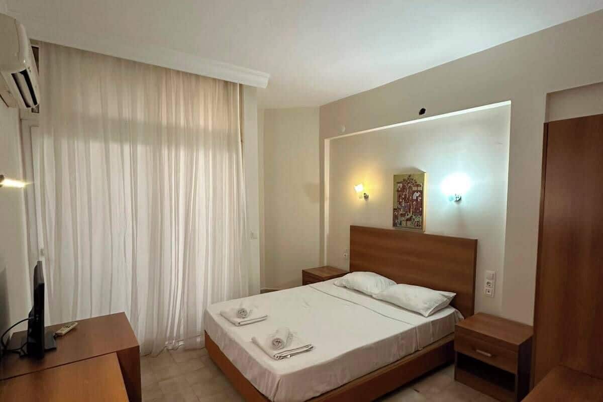 A hotel room with a bed, bedside tables, a desk, an air conditioning unit, and a window with curtains.