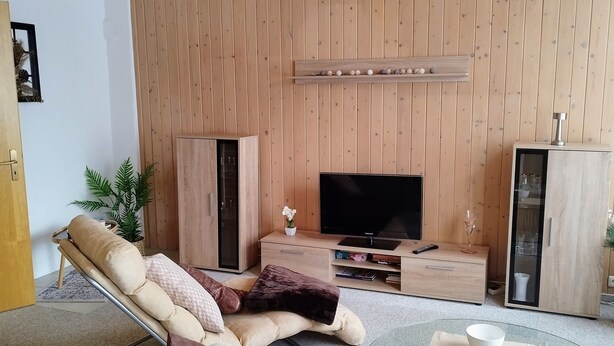 A modern living room with a wooden panel wall, a flat-screen TV, a glass coffee table, and a beige sofa. - Beautiful bright 60 sqm apartment near Magdeburg city center and zoo, barrier-free (Magdeburg)