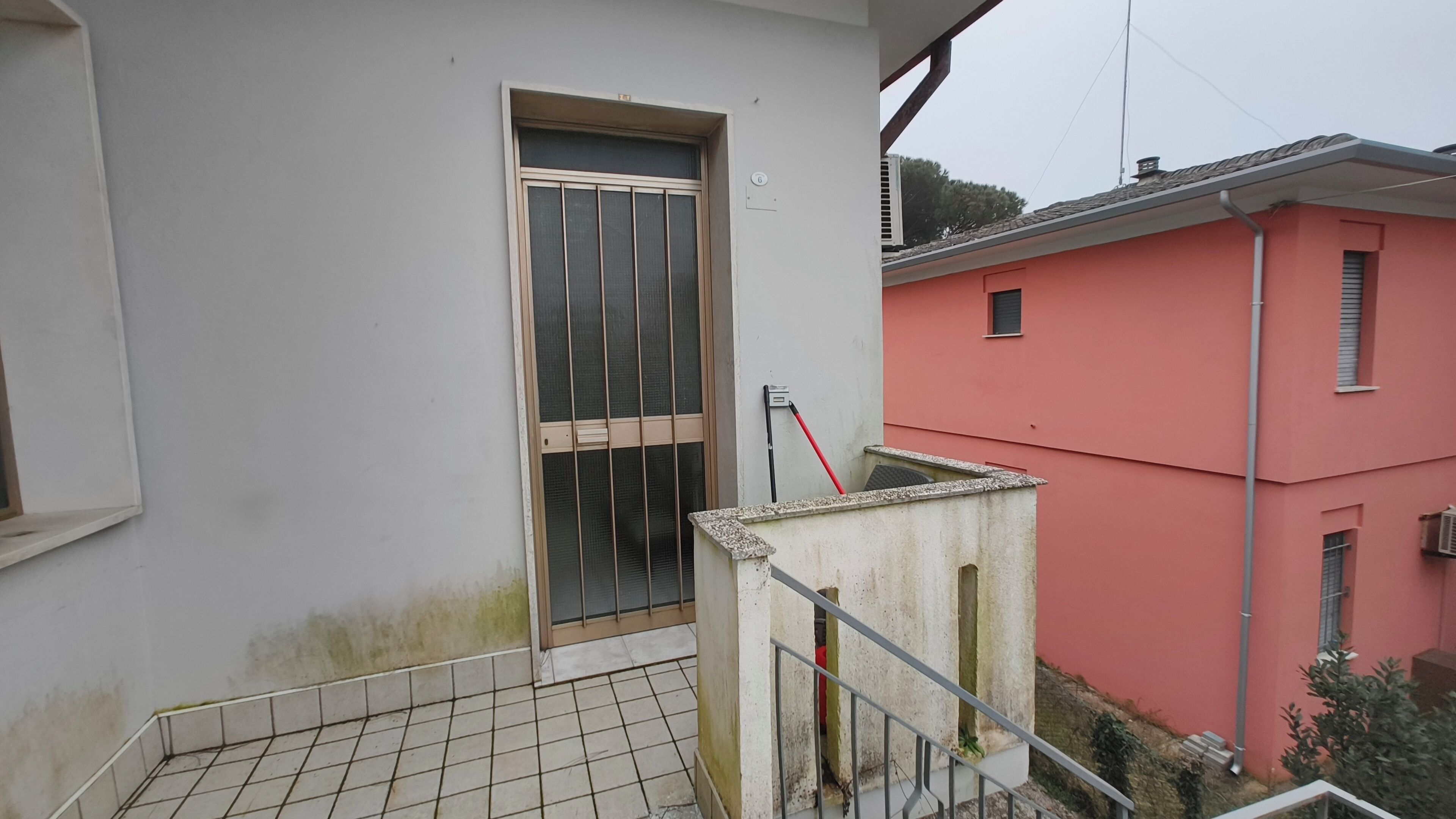 A building with a barred window and a red door, a tiled floor, and a metal railing.