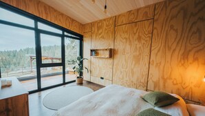 A modern bedroom with a large window, wooden walls, a bed with pillows, a bedside table, and a small plant.